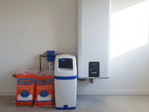 Assemblage de composants de plomberie disposés sur fond blanc : raccords en PVC blanc coudés, coudes en cuivre, raccords laiton, bonde d'évacuation chromée avec grille, joint d'étanchéité blanc, vis de fixation